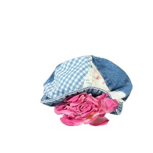 May include: A denim newsboy cap with a blue and white gingham pattern and floral patchwork. A large, dried pink rose rests beneath the brim. The hat is a unique accessory.