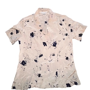 May include: A cream-colored button-down shirt with short sleeves. The shirt is decorated with a pattern of dark blue floral designs and thin stems. The fabric has a slight sheen, and the shirt has a collar.