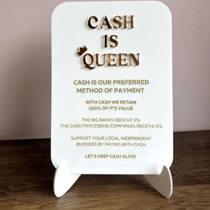 May include: White sign with gold lettering that reads "CASH IS QUEEN". The sign also includes the text "CASH IS OUR PREFERRED METHOD OF PAYMENT. WITH CASH WE RETAIN 100% OF IT'S VALUE. THE BIG BANKS RECEIVE 0%. THE CARD PROCESSING COMPANIES RECEIVE 0%. SUPPORT YOUR LOCAL INDEPENDENT BUSINESS BY PAYING WITH CASH. LET'S KEEP CASH ALIVE!"