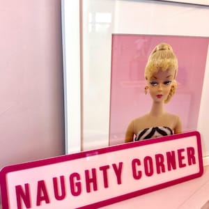 May include: A pink sign with a white border and the text "NAUGHTY CORNER" in white letters. The sign is in front of a framed picture of a blonde doll with a pink background.