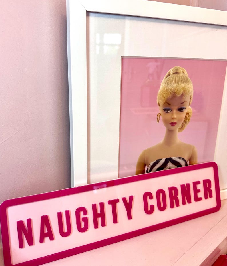 May include: A pink sign with a white border and the text "NAUGHTY CORNER" in white letters. The sign is in front of a framed picture of a blonde doll with a pink background.