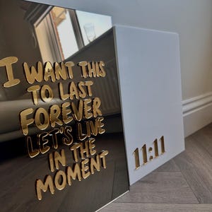 May include: Two decorative wall art pieces. One is a mirrored surface with gold lettering that reads "I WANT THIS TO LAST FOREVER LET'S LIVE IN THE MOMENT." The other is white with gold "11:11" lettering.