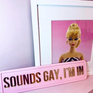 May include: Pink sign with gold lettering that reads "SOUNDS GAY, I'M IN". A framed picture of a blonde doll with a pink background is in the background.
