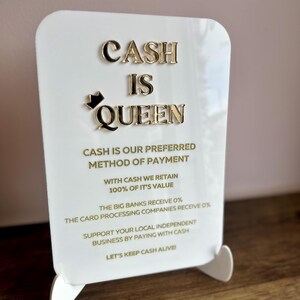 May include: White sign with gold lettering that reads "Cash is Queen". The sign also includes the text "Cash is our preferred method of payment. With cash we retain 100% of it's value. The big banks receive 0%. The card processing companies receive 0%. Support your local independent business by paying with cash. Let's keep cash alive."