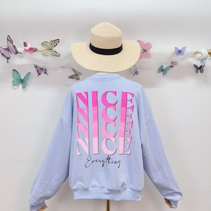 Może przedstawiać: Jasnoniebieska bluza z napisem "NICE" w różowym gradiencie powtórzonym cztery razy na plecach, z napisem "Everything" poniżej kursywą. Prezentowany jest kapelusz ze słomy z czarną wstążką.