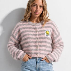 Könnte beinhalten: Eine flauschige, langärmelige Strickjacke mit Knopfleiste und horizontalen Streifen in Hellrosa und Grau. Der Strickpullover hat einen Rundhalsausschnitt und einen kleinen rechteckigen Aufnäher auf der linken Brust mit einem gelben Smiley-Symbol und Text.