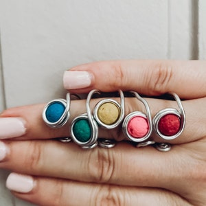 Könnte beinhalten: Fünf silberne Ringe mit bunten runden Perlen. Die Perlen sind türkis, grün, gelb, rosa und rot.