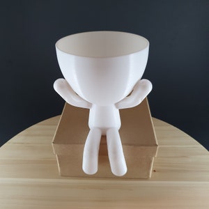 May include: White ceramic planter shaped like a person sitting with arms outstretched. The planter has a round top for planting.