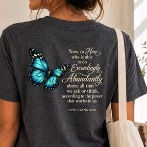 May include: A dark gray short-sleeve t-shirt featuring a blue butterfly graphic and the text: Now to Him who is able to do Exceedingly Abundantly above all that we ask or think, according to the power that works in us. EPHESIANS 3:20.