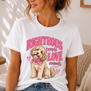 May include: A white short-sleeved t-shirt featuring a graphic of a light brown fluffy dog wearing a pink heart-patterned bandana. The text reads RIGHTEOUS people LOVE animals PROVERBS 12:10, accompanied by two pink heart outlines on the sides.
