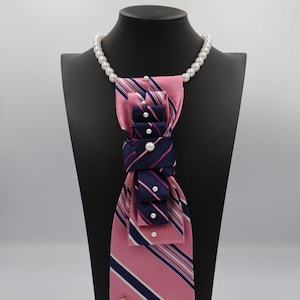 May include: A necktie accessory featuring a pink and navy blue striped fabric with a knotted design. The piece is adorned with white faux pearls throughout the fabric and a string of white pearls around the neck. A gold safety pin with blue beads is attached.