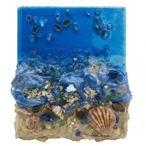 Può includere: Un'opera d'arte in resina blu e bianca che rappresenta un paesaggio marino con una spiaggia sabbiosa, onde blu e conchiglie. Il pezzo è realizzato con vetro e resina incorporati.