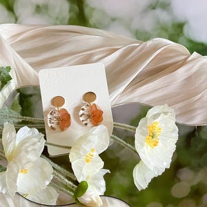 Peut inclure: Boucles d'oreilles artisanales avec des clous dorés et des pendentifs en résine transparente contenant des fleurs orange séchées et des détails blancs. Le texte "West Moon" est visible sur la carte.