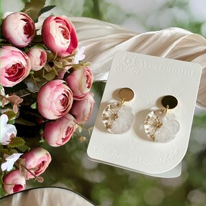 Peut inclure: Boucles d'oreilles rondes transparentes avec des inclusions florales dorées et blanches, surmontées d'un disque doré. Elles sont présentées sur une carte blanche avec l'inscription "Westmount" et "Gift from garden". Des roses roses en arrière-plan.