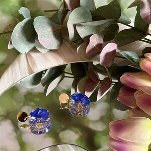 Peut inclure: Une paire de boucles d'oreilles bleues et dorées avec un motif floral. Les boucles d'oreilles sont rondes et ont une tige dorée. Les boucles d'oreilles sont présentées sur une surface réfléchissante avec de la verdure et des fleurs en arrière-plan.