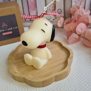 May include: A Snoopy-shaped candle with a red collar and black ears, sitting on a wooden tray. A red and white checked ribbon is tied to the top of the candle. The Little Candle Lab logo is visible on a brown box in the background.