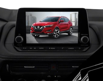 May include: A red Nissan Kicks SUV is displayed on the screen of a car's infotainment system. The screen shows the car's exterior, with the front of the car facing the camera. The screen is surrounded by black plastic trim and buttons. The buttons include "AUDIO", "MENU", "CAMERA", and "PUSH SOUND".