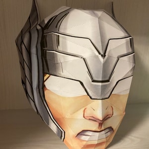 May include: A white papercraft mask of Thor, a superhero from Marvel Comics. The mask is detailed with a textured, geometric design and features a fierce expression.