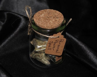 May include: A small glass jar with a cork lid and a brown paper tag that reads "Personal Power Spell". The jar is filled with dried herbs.