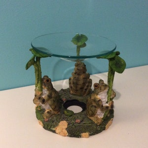 May include: A glass oil burner with a round glass top and three frogs sitting on a green base. The frogs are holding lily pads.