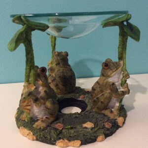K&ouml;nnte beinhalten: Ein Keramik-Frosch-&Ouml;lbrenner mit drei Fr&ouml;schen, die auf einer gr&uuml;nen Basis sitzen. Die Fr&ouml;sche sind braun und gr&uuml;n mit schwarzen Streifen. Der &Ouml;lbrenner hat eine klare Glasschale oben.