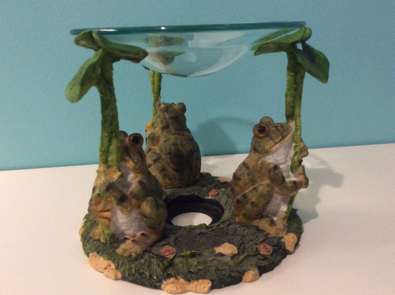 K&ouml;nnte beinhalten: Ein Keramik-Frosch-&Ouml;lbrenner mit drei Fr&ouml;schen, die auf einer gr&uuml;nen Basis sitzen. Die Fr&ouml;sche sind braun und gr&uuml;n mit schwarzen Streifen. Der &Ouml;lbrenner hat eine klare Glasschale oben.
