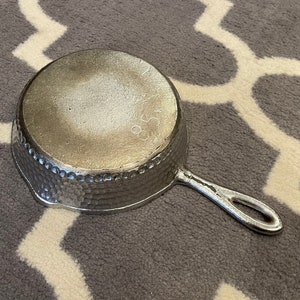 Op de afbeelding: Een zilveren gehamerde gietijzeren koekenpan met een handvat. De koekenpan heeft een getextureerd oppervlak en het nummer 8,5 is zichtbaar op de bodem.