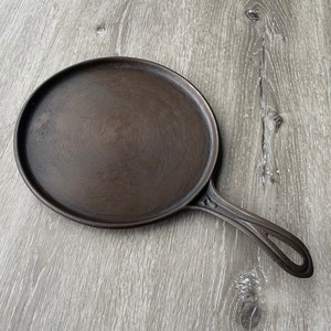 May include: A cast iron pizza pan with a dark brown patina. The pan has a round cooking surface and a long handle.