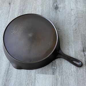 May include: A cast iron skillet with a long handle and a circular cooking surface. The underside of the pan is visible, with the text "Wagner Ware Sidney -O-" and the number "8" embossed. The pan is dark gray and sits on a light gray wooden surface.