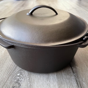 Op de afbeelding: Een zwarte gietijzeren stoofpot met een deksel en twee handvatten. De stoofpot staat op een houten oppervlak.