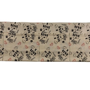 Op de afbeelding: Een beige stoffen deurstopper met een hondenmotief met de tekst "Ik hou van honden" die in zwarte letters wordt herhaald. De deurstopper heeft aan beide uiteinden twee metalen ringen.