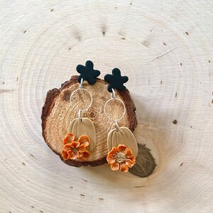 Peut inclure: Boucles d'oreilles faites à la main avec un clou en forme de fleur noire, un cercle argenté, une citrouille beige et une fleur orange. Présentées sur une tranche de bois, parfaites pour l'automne.