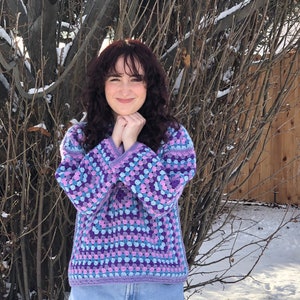Puede incluir: Un suéter de ganchillo hecho a mano con un patrón de cuadrados de la abuela. El suéter presenta una paleta de colores de cuadrados morados, azules y lavanda. Las mangas y el cuello están ribeteados con un borde liso de lavanda. La persona lleva vaqueros azules.