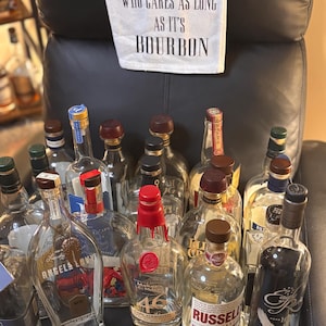 Peut inclure: Plusieurs bouteilles de bourbon vides avec des étiquettes et des formes variées. En arrière-plan, un tissu blanc porte l'inscription "GLASS HALF EMPTY OR GLASS HALF FULL WHO CARES AS LONG AS IT'S BOURBON".