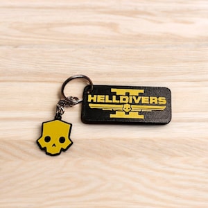 Könnte beinhalten: Ein schwarzer Leder-Schlüsselanhänger mit dem Text "HELLDIVERS" in gelben Buchstaben. Der Schlüsselanhänger hat einen gelben Totenkopf-Anhänger.