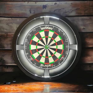Pode incluir: Um alvo de dardos Winmau Blade 6 com uma moldura preta e iluminação integrada. O alvo de dardos apresenta um design clássico com segmentos vermelhos, verdes, pretos e brancos. Os números são claramente visíveis.