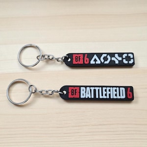 Könnte beinhalten: Zwei schwarze Schlüsselanhänger mit silbernen Schlüsselringen und Ketten. Ein Schlüsselanhänger hat den Text "BF 6" in Rot und weißen Symbolen. Der andere Schlüsselanhänger hat den Text "BF BATTLEFIELD 6" in Rot und Weiß.