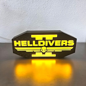 Könnte beinhalten: Ein beleuchtetes, schwarzes Schild mit dem gelben Schriftzug "HELLDIVERS" und einem Totenkopf-Flügel-Design. Das rechteckige Schild mit abgeschrägten Kanten strahlt warmes, gelbes Licht aus.