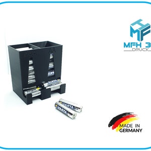 Könnte beinhalten: Schwarzer Batterie-Organizer aus Kunststoff mit zwei Fächern, die mit "AAA" und "AA" beschriftet sind. Der Organizer ist mit Batterien verschiedener Marken gefüllt. Zwei VARTA-Batterien liegen auf einer weißen Oberfläche vor dem Organizer.