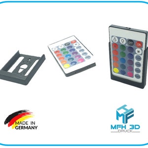 Puede incluir: Un mando a distancia de plástico negro con 16 botones, que incluyen opciones de color, fundido y velocidad. El mando a distancia está en un soporte de plástico negro. También se muestra un soporte de plástico negro para una bombilla. El texto "Made in Germany" es visible en la imagen.