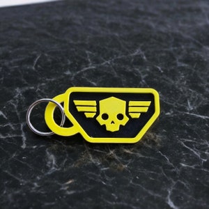 Pode incluir: Um chaveiro amarelo com um design de caveira e asas pretas. O chaveiro tem um anel prateado.