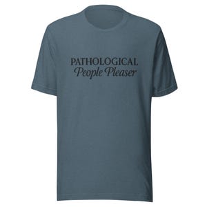 Könnte beinhalten: Ein kurzärmeliges, teal-farbenes T-Shirt mit dem schwarzen Schriftzug "PATHOLOGICAL People Pleaser" in Schreibschrift auf der Brust. Das Shirt hat einen Rundhalsausschnitt und eine lockere Passform.