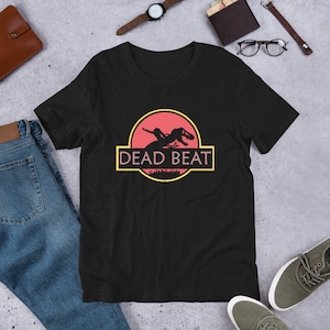 Pode incluir: Camiseta preta com um gráfico vermelho e amarelo de um dinossauro com o texto "DEAD BEAT" impresso nela.