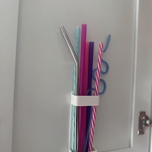 Puede incluir: Un soporte blanco de plástico para pajitas montado en una puerta de armario blanca, lleno de una variedad de pajitas de colores. Las pajitas incluyen una pajita de metal plateado, pajitas rosas, moradas, azules y a rayas. El soporte es rectangular con dos bandas.
