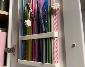 May include: A variety of reusable straws in a white organizer mounted inside a cabinet. The straws are in various colors, including pink, green, blue, and clear. Some straws have decorative flower accents.