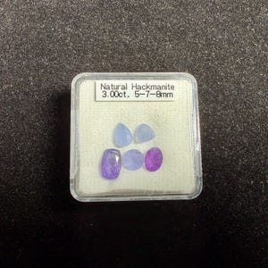 May include: A clear plastic case contains five natural hackmanite gemstones. The gemstones are various shapes, including oval and teardrop, and range in color from light blue to purple. The label reads "Natural Hackmanite 3.00ct. 5-7-8mm."