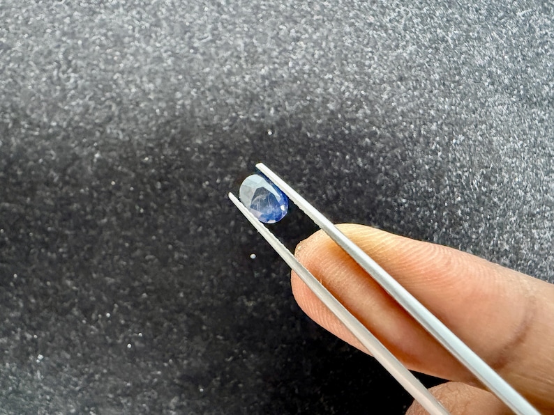 May include: A small, oval-shaped, deep blue gemstone held by a pair of silver-toned metal tweezers. The stone features a smooth, polished surface with visible internal facets and a translucent quality, reflecting light across its dark blue hue.
