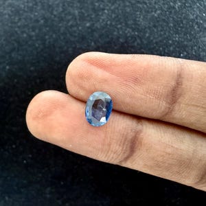 May include: An oval-cut blue gemstone with a faceted surface that reflects light. The stone displays a translucent appearance with varying shades of deep and light blue throughout its structure, showcasing a polished finish and a clear, brilliant clarity.