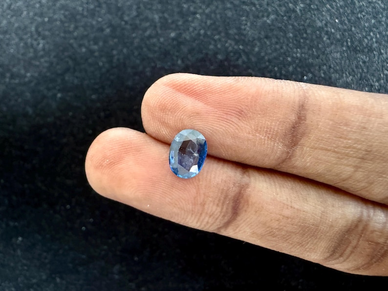 May include: An oval-cut blue gemstone with a faceted surface that reflects light. The stone displays a translucent appearance with varying shades of deep and light blue throughout its structure, showcasing a polished finish and a clear, brilliant clarity.