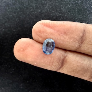 May include: An oval-cut gemstone with a faceted surface and a translucent blue color. The stone displays varying shades of light and dark blue throughout its structure, reflecting light across its polished, angular surfaces.